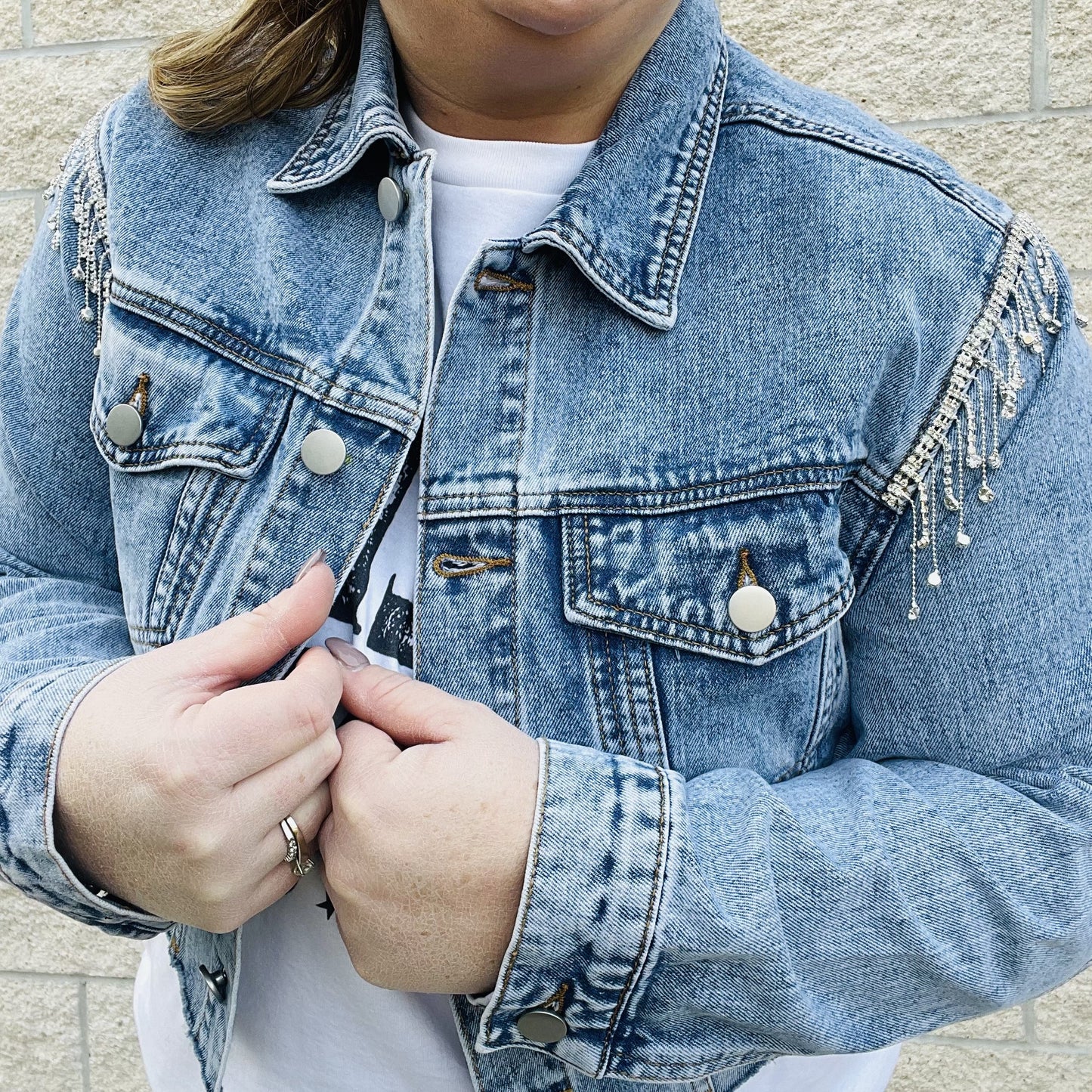 Elevate your style with this edgy denim jacket. With rhinestone details on the sleeves, this jacket will turn heads wherever you go. The distressed hemline gives it a unique edge that you'll love. Perfect for a night out or for everyday wear, this jacket is a must-have in your wardrobe.  Details Rhinestones details on sleeves Cropped Distress hemline 87% Cotton | 8% Poly | 5% Viscose
